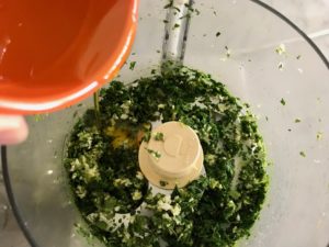 Food processor with garlic and fresh herbs and olive oil being poured in