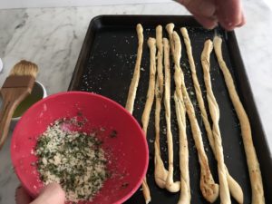 breadsticks on a baking sheet being sprinkled with the herb mixture
