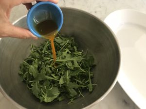 arugula in a mixing bowl with sun dried tomato oil being poured over