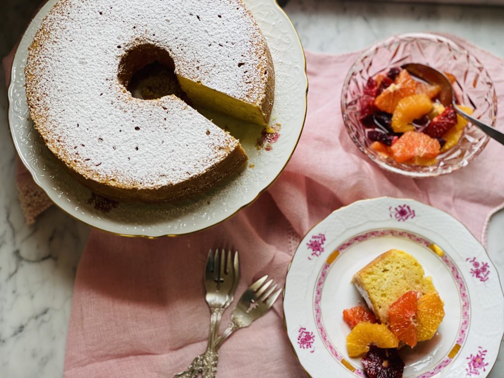 Round cake topped with white powdered sugar. Slice of cake on a nearby plate is topped with yellow, orange and purple citrus wedges.